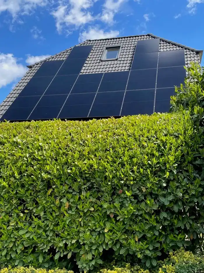 Hausdach mit grauen Ziegeln, Solarmodulen, einem Dachfenster und einem grünen Busch im Vordergrund unter blauem Himmel.
