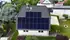 Dach eines weißen Hauses mit schwarzen Solarmodulen, umgeben von grünen Bäumen und Gärten.
