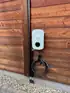 Weiße Ladestation mit Kabel und Stecker, montiert an einer braunen Holzfassade, davor Kies.